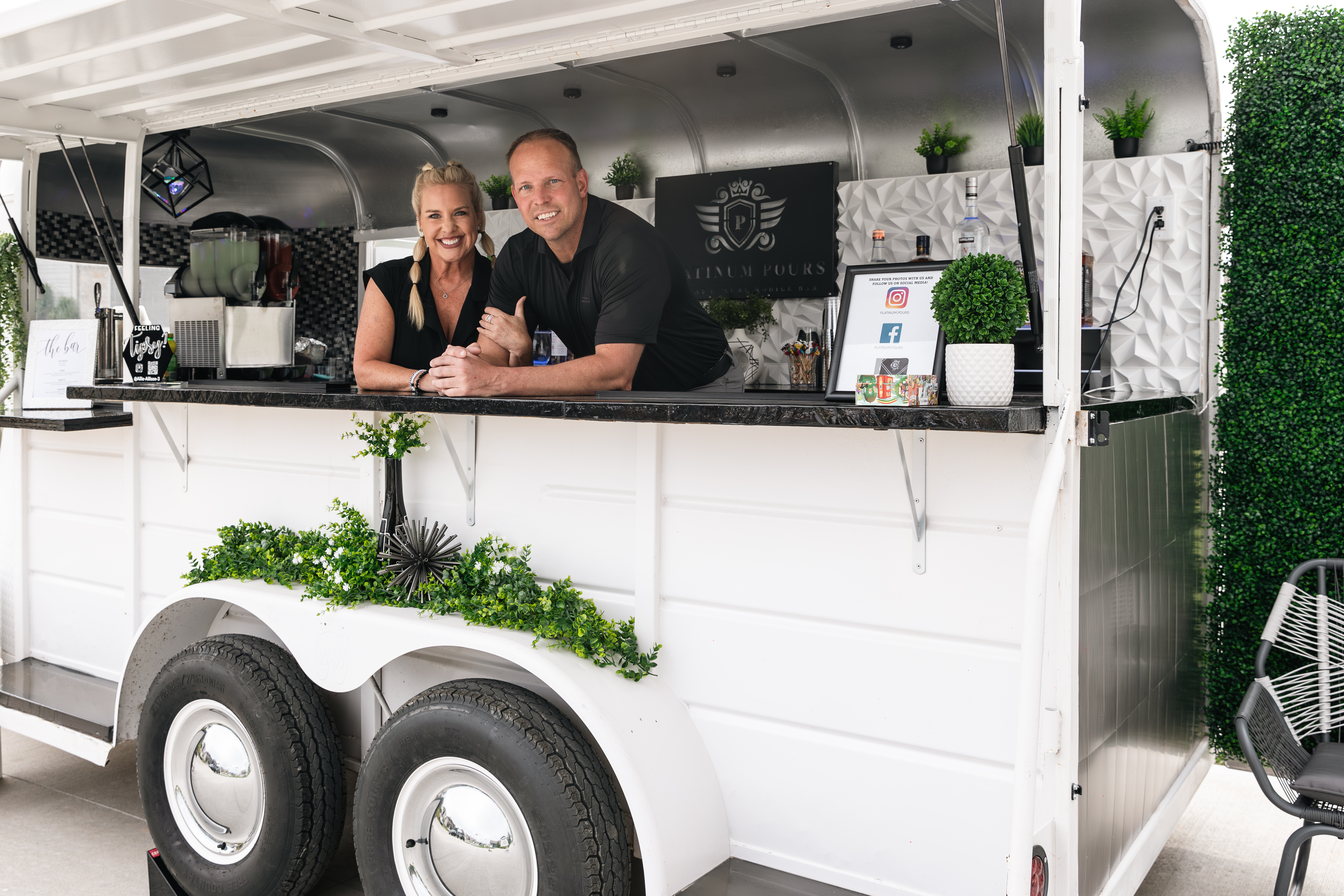 Scott and Allie, owners of Platinum Pours, smiling together behind the bar of Jackpot the luxury mobile trailer with the Platinum Pours crest backdrop, 3D LED tile and greenery visible inside