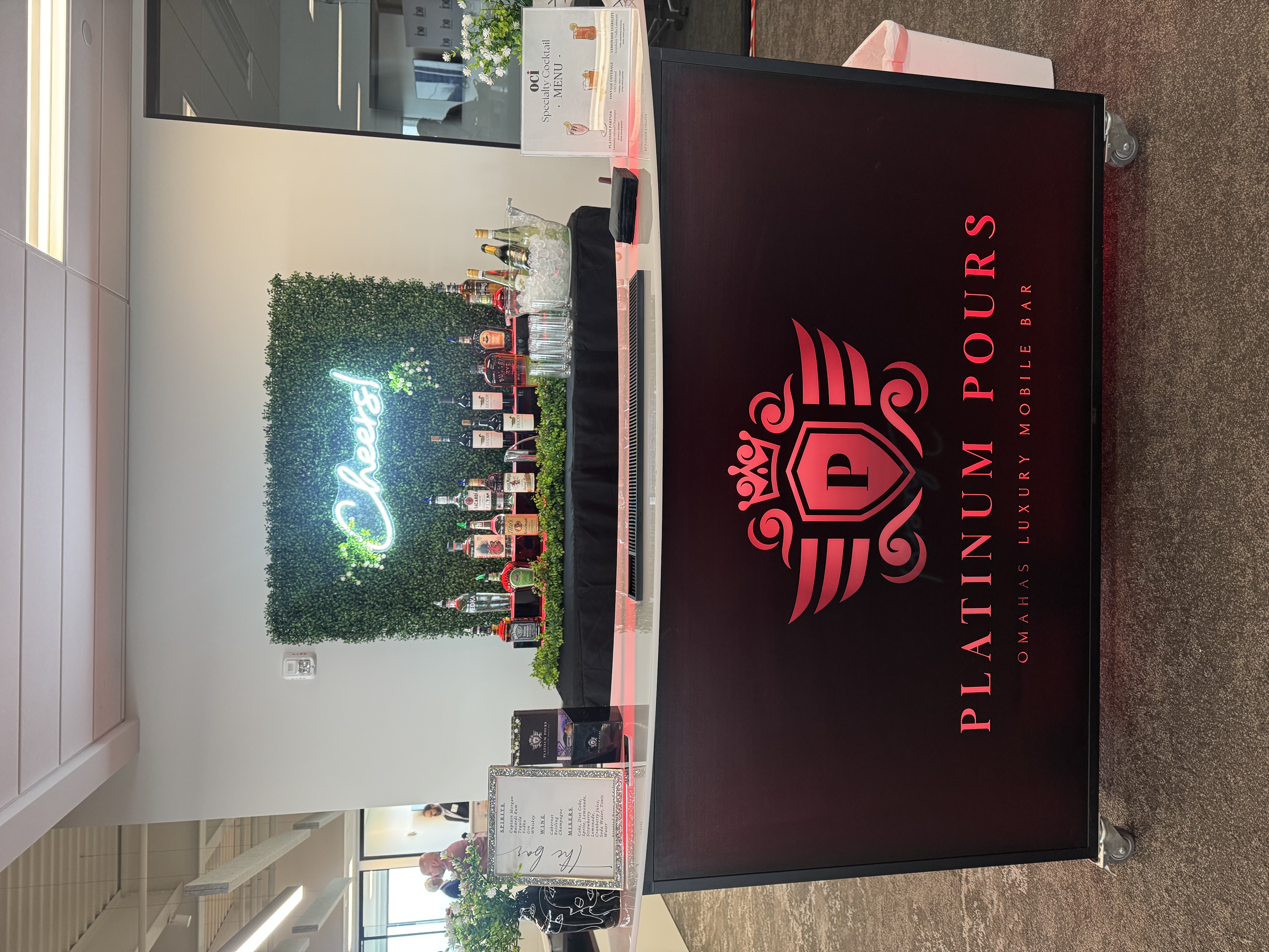 The Platinum Pours 6 ft LED rolling bar with the full crown and wing crest on the front, set up at a corporate event with the Cheers neon greenery wall, full bottle display, branded menu cards and floral styling behind it