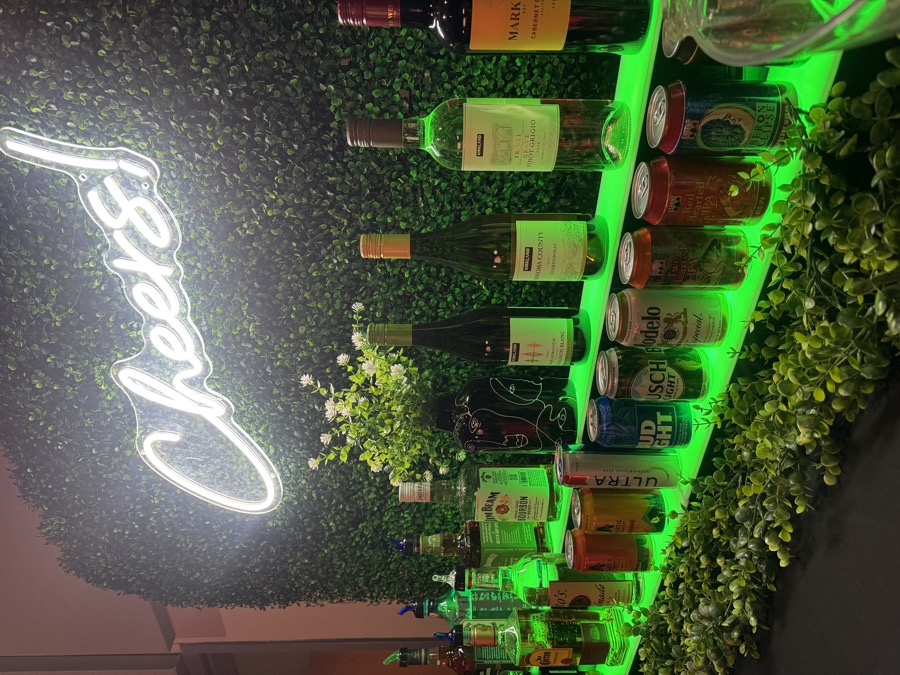 Curated wine, beer and spirits display lit in soft green against the Cheers boxwood wall