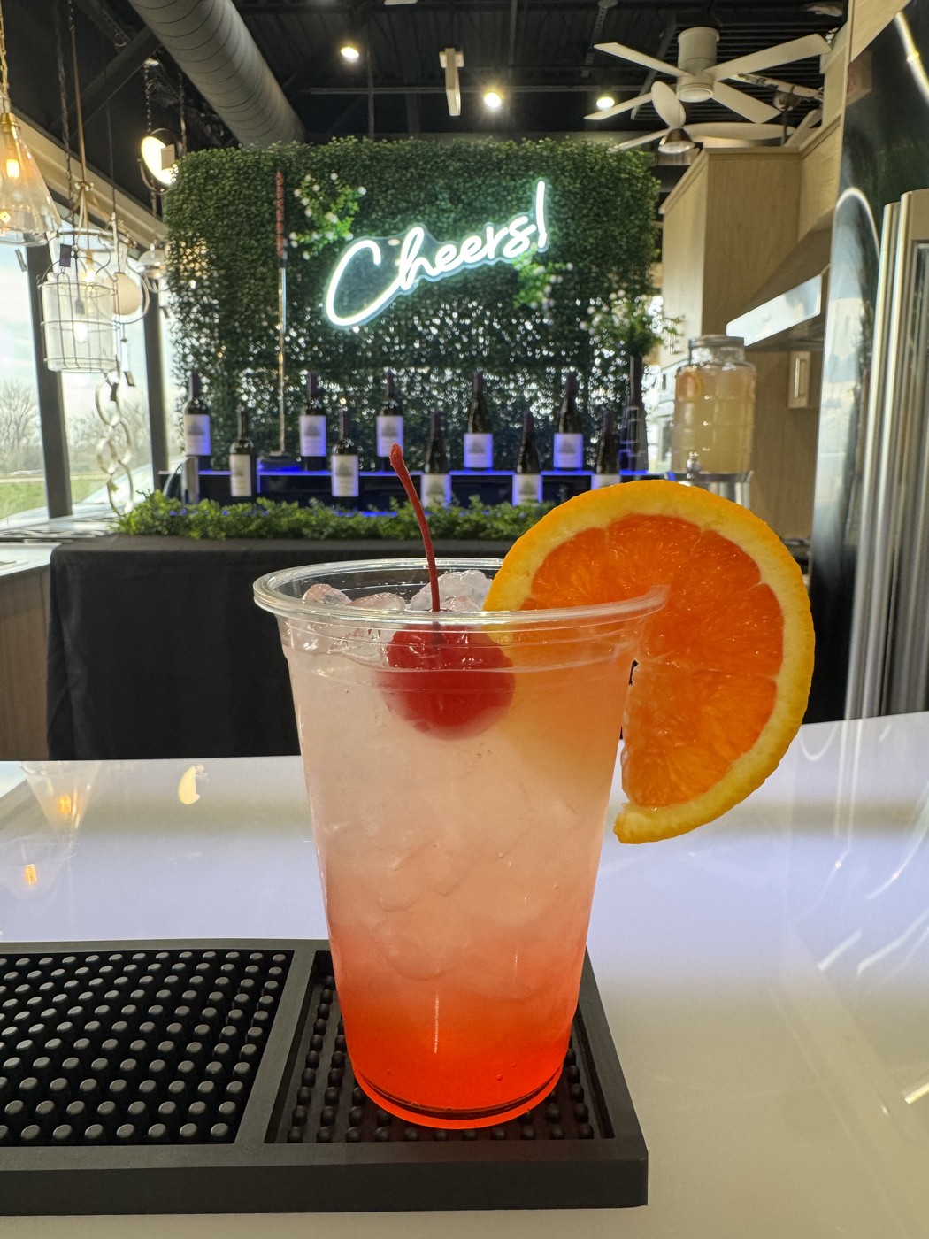 A signature Platinum Pours cocktail with orange wheel and cherry garnish, in front of the Cheers neon wall and wine display