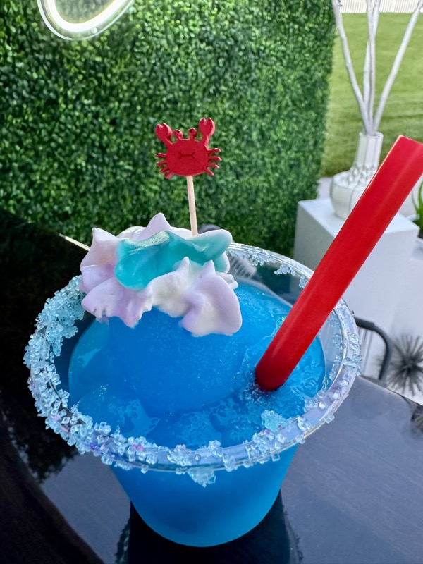 Blue frozen mocktail with whipped cream, gummy garnish and red candy straw, topped with a crab stirrer in front of the Cheers wall
