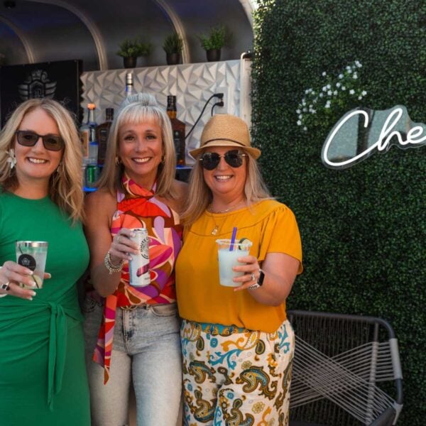 Three guests raising their glasses in celebration at a Platinum Pours event, with the mobile bar trailer, decorative ivy, and a neon sign in the background.